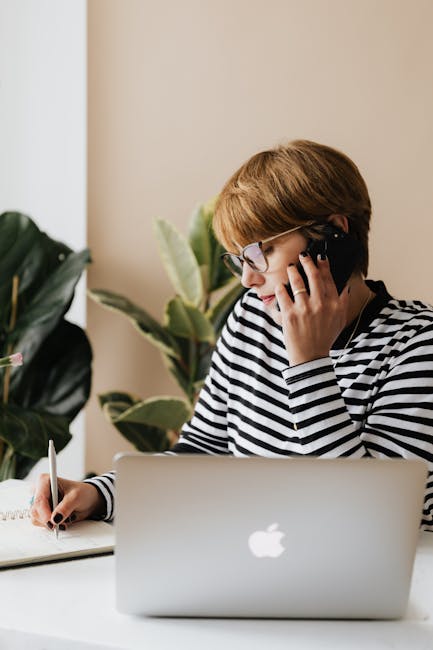 Concentrated female freelancer wearing casual clothes and eyeglasses sitting at table and having conversation on smartphone and writing notes in notepad whil…