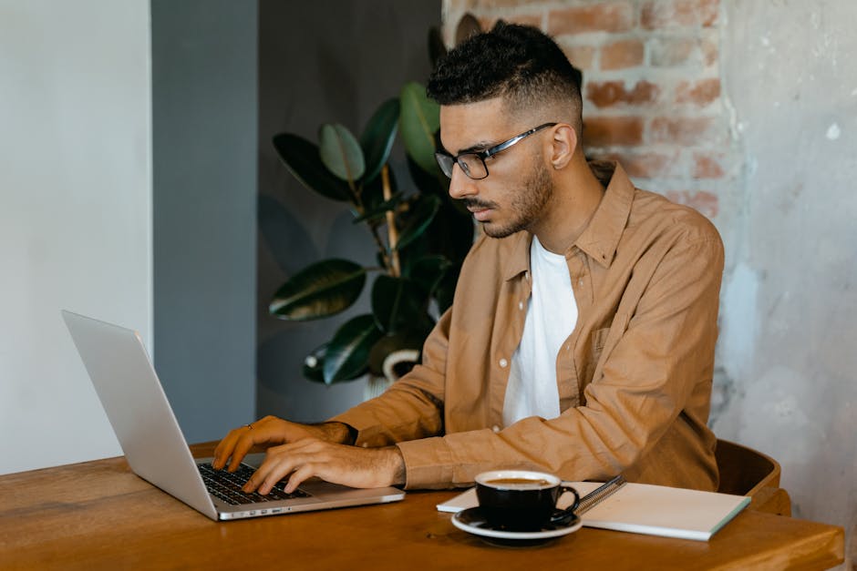 Young man with glasses working intently on laptop at a cafe.